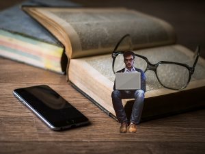 Image d'un livre, de lunettes et teléphone et un petit homme asis sur le livre un ordinateur sur les genoux