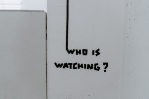 Tag mural who is watching ? en noir sur mur blanc. 