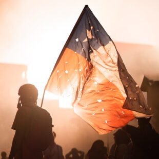 Photographie en paysage et en couleurs d'une scène de manifestation au Chili. Le premier plan montre un manifestant portant le drapeau chilien cribblé d'impacts de balles. La prise de vue est en lègère contre-plongée, laissant apparaître le ciel dans lequel se détache le drapeau au centre de l'image, et une lumière laissant supposer la présence de fumigènes. La foule apparaît au second plan.