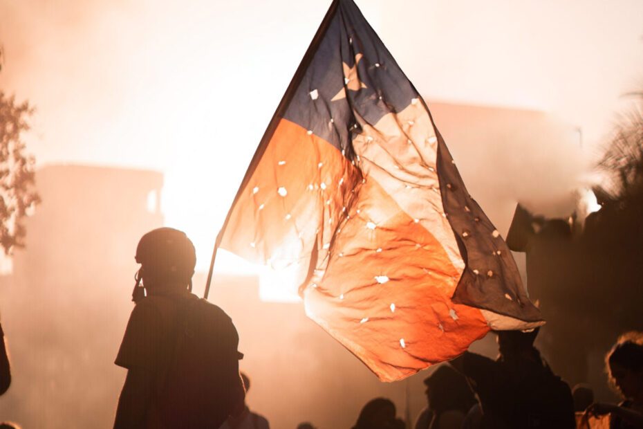 Photographie en paysage et en couleurs d'une scène de manifestation au Chili. Le premier plan montre un manifestant portant le drapeau chilien cribblé d'impacts de balles. La prise de vue est en lègère contre-plongée, laissant apparaître le ciel dans lequel se détache le drapeau au centre de l'image, et une lumière laissant supposer la présence de fumigènes. La foule apparaît au second plan.